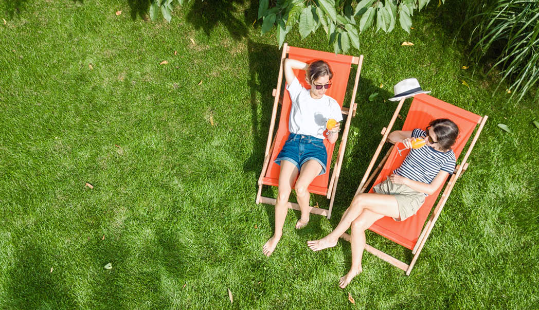 A top‑down view of two people relaxing on orange deck chairs on a lush green lawn, enjoying drinks and surrounded by leafy garden plants.